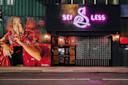 A street view of a building at night with a large mural of a woman in a red dress holding a finger to her lips, next to a neon sign reading "SEI LESS" above a black entrance.
