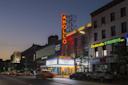 The Apollo Theater at dusk, with its bright vertical neon sign and marquee lit up. Nearby, there are storefronts for Banana Republic, GameStop, and Cinderella Eyebrows, with cars parked along the street.