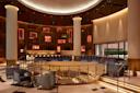 Luxurious hotel lobby with a circular design, featuring a central staircase, large columns, and ambient lighting. Modern seating with patterned chairs surrounds the area. Artwork is displayed on a wooden wall with illuminated frames.