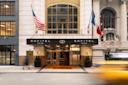 Entrance of Sofitel New York, featuring a modern facade with two flags, one American and one French. A blurred yellow taxi passes by on the street, capturing a dynamic city scene.