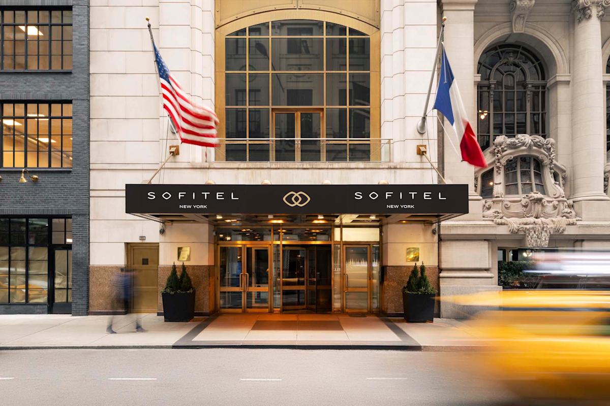 Entrance of Sofitel New York, featuring a modern facade with two flags, one American and one French. A blurred yellow taxi passes by on the street, capturing a dynamic city scene.