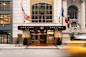 Entrance of Sofitel New York, featuring a modern facade with two flags, one American and one French. A blurred yellow taxi passes by on the street, capturing a dynamic city scene.