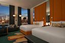 Modern hotel room with two queen beds, orange headboards, a dark bench, sofa, and large windows offering a city skyline view at dusk. Contemporary decor includes patterned rug and decorative pillows.