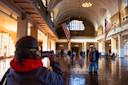 A person in a red hoodie takes a photo of two people posing inside a large, sunlit hall with arched windows, high ceilings, and two large American flags hanging from the ceiling. Other visitors are seen in the background.