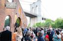 A large crowd gathers outside a brick building with an arched entrance under the Brooklyn Bridge. People are talking, holding drinks, and socializing at an outdoor event near a sign reading "STUDIO.