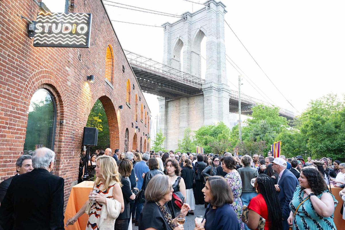 A large crowd gathers outside a brick building with an arched entrance under the Brooklyn Bridge. People are talking, holding drinks, and socializing at an outdoor event near a sign reading "STUDIO.