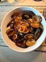 A white bowl filled with seafood pasta, including clams, mussels, calamari, and pasta in a rich tomato sauce, sits on a rustic white wooden table.