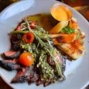 A plated meal featuring grilled steak topped with chimichurri, cherry tomatoes, and rosemary, served with seasoned potato wedges and a small cup of creamy dipping sauce.