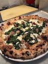 A close-up of a wood-fired pizza topped with broccoli rabe, crumbled sausage, and melted cheese, served on a white plate on a marble counter.