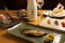 A Japanese meal with grilled fish on a green plate, assorted sashimi, tempura, and sushi, served with a bottle of sake and a glass on a wooden table.