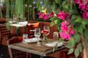A table set for two with wine glasses, plates, and a small vase of red flowers, surrounded by lush pink and green foliage in an outdoor restaurant setting.