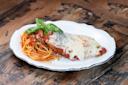 A white plate with spaghetti topped with tomato sauce and fresh basil, next to a breaded chicken cutlet covered in melted cheese and tomato sauce, served on a rustic wooden table.