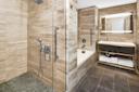 Modern bathroom with beige marble tiles features a spacious shower with glass doors, a bathtub, and a vanity with a large mirror. Towels are neatly stacked on the shelf beneath the sink. The floor is tiled in dark gray.