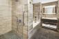 Modern bathroom with beige marble tiles features a spacious shower with glass doors, a bathtub, and a vanity with a large mirror. Towels are neatly stacked on the shelf beneath the sink. The floor is tiled in dark gray.