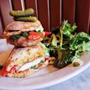 A ciabatta sandwich with grilled chicken, spinach, and red peppers, topped with two pickles on a skewer, served with a side of mixed green salad on a white plate.