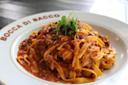 A plate of tagliatelle pasta with a rich, meaty tomato sauce, garnished with fresh greens. The dish is served on a white plate with "Bocca di Bacco" written on the rim.