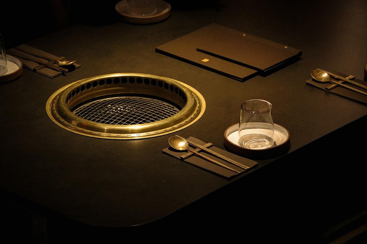 A dimly lit table set for dining features a built-in grill, utensils, plates, and glasses neatly arranged, creating an inviting atmosphere for a meal.