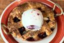 A small berry pie with a golden lattice crust sits in a round dish, topped with a scoop of vanilla ice cream with a swirl of red fruit sauce, on a red and white plate.