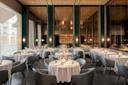 Elegant restaurant interior with round tables covered in white tablecloths, surrounded by modern grey chairs; soft lighting, mirrored walls, and a stylish bar area in the background create a sophisticated atmosphere.