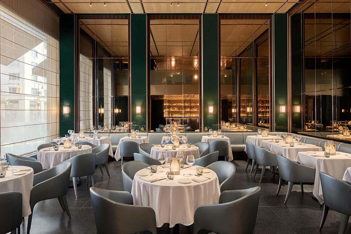 Elegant restaurant interior with round tables covered in white tablecloths, surrounded by modern grey chairs; soft lighting, mirrored walls, and a stylish bar area in the background create a sophisticated atmosphere.