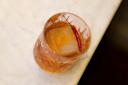 A glass filled with a brown cocktail garnished with a red chili pepper and a large ice cube, placed on a marble surface.