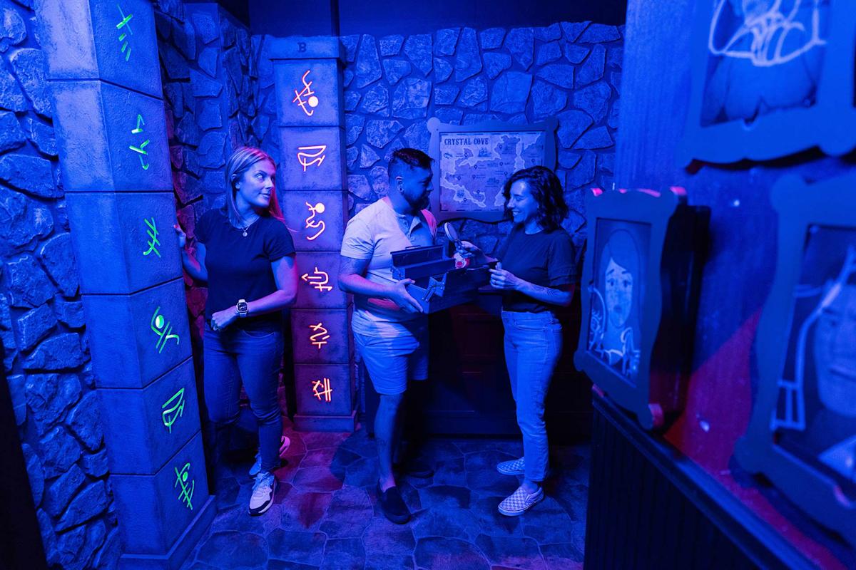 Three people stand in a dimly lit, neon-lit room with stone walls and mysterious glowing symbols, appearing to solve a puzzle together, likely in an escape room setting.
