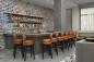 Modern bar interior with a long wooden counter and tan leather stools lined up along it. Behind the bar, shelves hold various bottles. The back wall features a colorful tiled mosaic, and natural light filters through a window with sheer curtains.