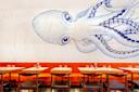 A dining area with rows of wooden tables and dark chairs against a wall featuring a large, artistic blue and white octopus mural. The tables are set with glassware and napkins, and the seating includes orange cushions.