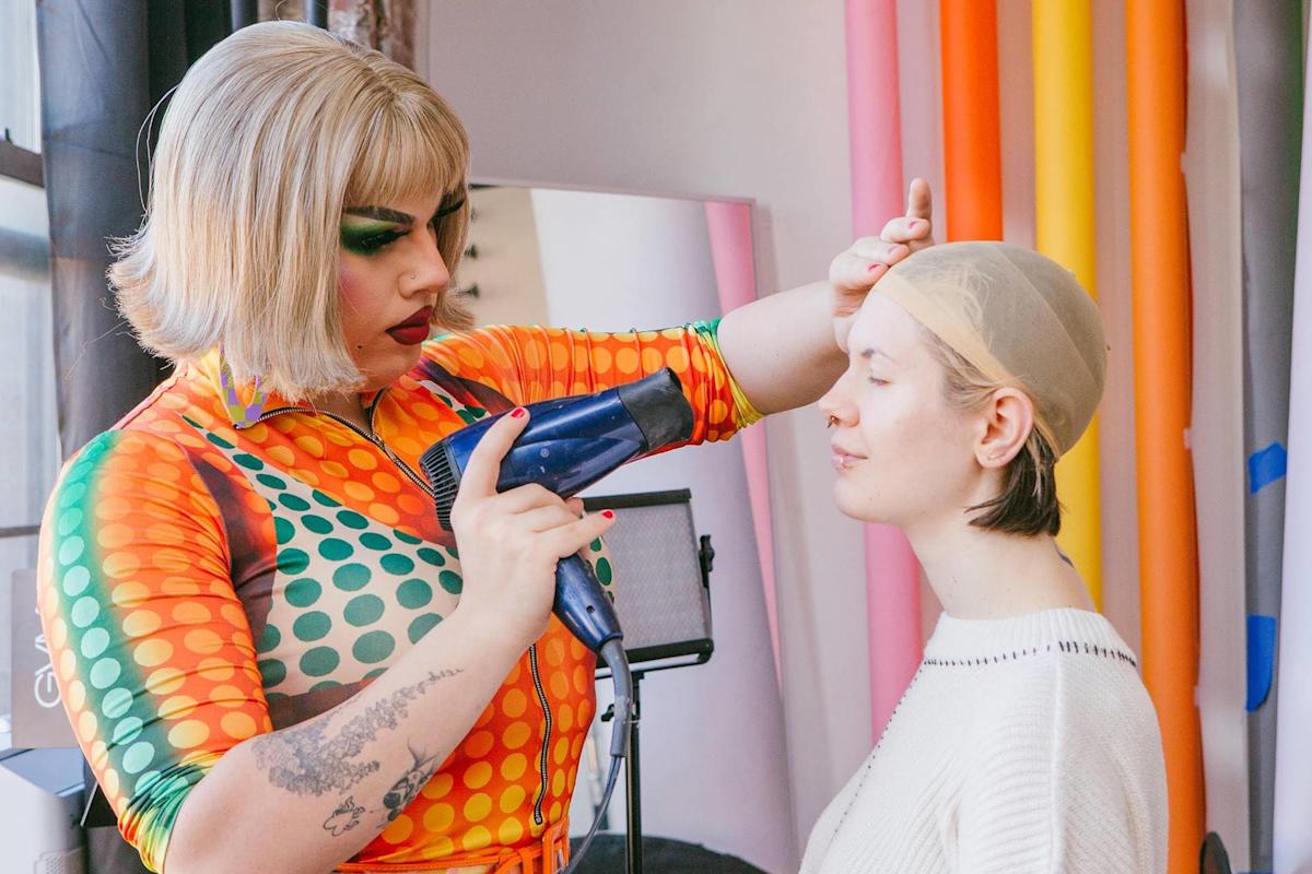A person in a colorful, polka-dot outfit uses a blow dryer on another person's head, who is wearing a wig cap and sitting with eyes closed, in a room with colorful backdrops.