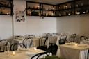 A cozy restaurant interior with white tablecloth-covered tables and black chairs. Candles are placed on tables, and glassware is set. Shelves with jars and decor line the walls, and a black and white photo is displayed on one wall.