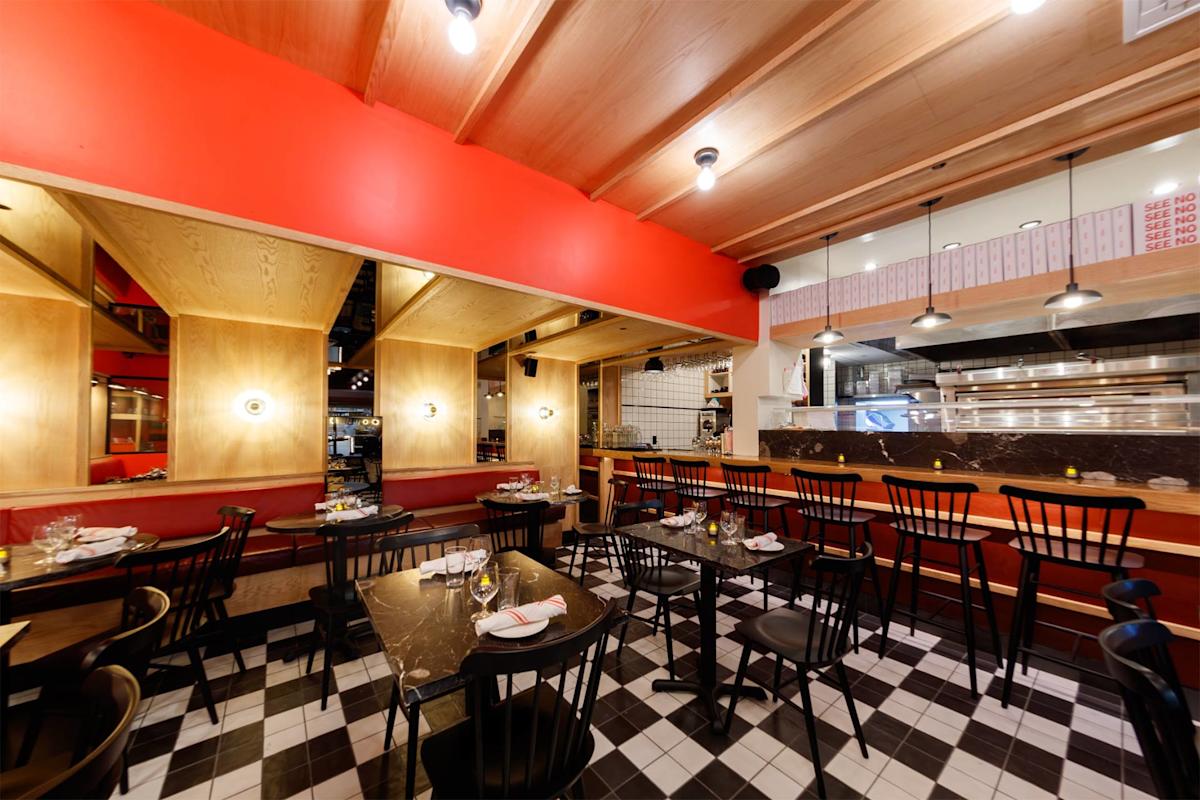 A modern restaurant interior with black chairs, dark tables, checkered black-and-white tile floor, red accent walls, wooden ceiling beams, and an open kitchen visible in the background. Tables are set with glasses and napkins.