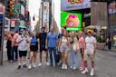 A group of nine people is posing on a busy city street, surrounded by tall buildings and bright advertisements. They are casually dressed and smiling. A large advertisement featuring a cartoon bear with a bow is visible in the background.