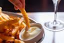 A hand dips a French fry into a small bowl of creamy white sauce next to a plate of fries, with a glass visible in the background.