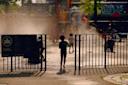 A child enters a playground where several children play in water mist on a sunny day. Sunlight creates reflections on the wet ground, and a playground sign and gate are visible in the foreground.