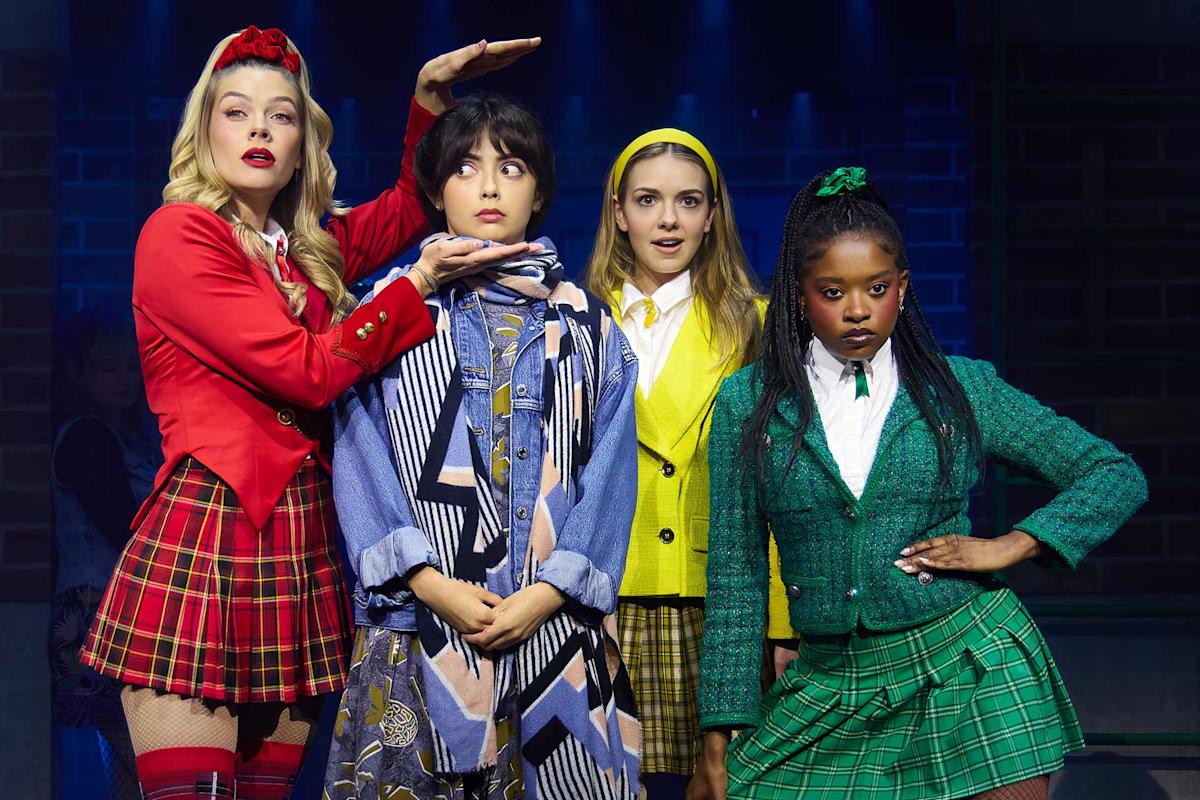 Four women in colorful, preppy outfits pose dramatically on stage. One in red gestures above another’s head, who looks surprised. Two others in yellow and green stand confidently beside them under stage lights.