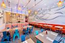 A modern restaurant interior featuring teal metal chairs, orange cushioned benches, and hanging woven light fixtures. The walls showcase a blue octopus mural. The bar at the back is stocked with bottles and has a hanging light system with red wires.