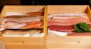Two wooden boxes filled with assorted raw fish fillets, including various cuts of tuna, salmon, and white fish, with a garnish of green shiso leaves on top of the fish in the right box.