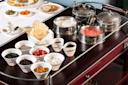 An assortment of ice cream toppings and sauces in small white bowls, with containers of ice cream and whipped cream on a serving cart; a table with plates and drinks is visible in the background.