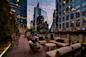 Rooftop patio at dusk with string lights, modern sofas, chairs, and tables. Surrounded by tall glass buildings, the space has a cozy atmosphere and cityscape views under a colorful evening sky.