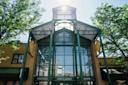 Glass entrance of Jamaica Market with green metal framework, trees on both sides, and sunlight shining from above. Signs read "Food Court," "Jamaica Market," and "Shops.