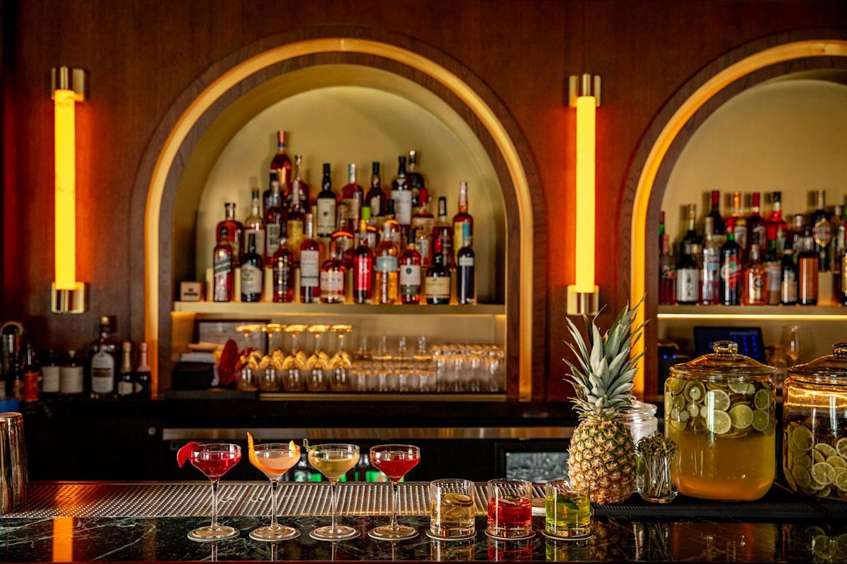 A bar with illuminated shelves displays various liquor bottles. In front, six colorful cocktails, a pineapple, and two large jars of infused drinks sit on the counter. Amber wall lights frame the arched shelves.