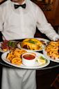 A server in a white shirt and black bow tie carries a tray with plates of curly fries, a baked potato topped with melted cheese, steak slices, and a bowl of marinara sauce with lemon wedges.