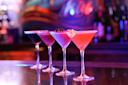 A row of four colorful martini glasses with different cocktails, each garnished distinctively, sits on a reflective bar counter. The background is blurred with vibrant lights and bottles, creating a lively atmosphere.