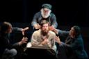 A bearded man sits center stage, looking contemplative, surrounded by three people. Two individuals, dressed in dark clothing, reach toward him, while an older man in a cap and jacket stands behind, also gesturing toward him. The scene is dramatic.