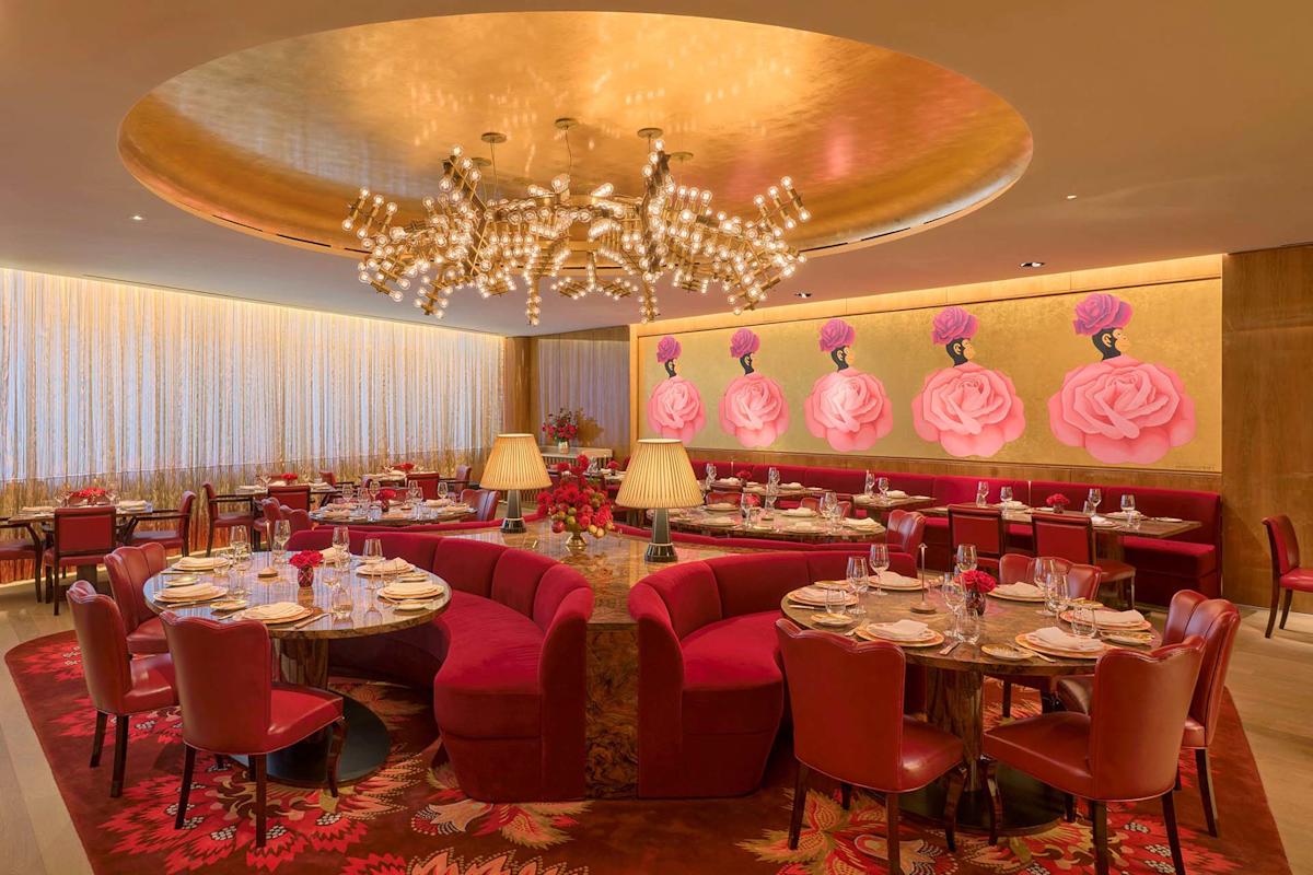 Elegant restaurant with round red velvet seating, gold ceiling, modern chandelier, and pink floral wall art. Tables are set with glassware and plates, creating a warm, luxurious atmosphere.