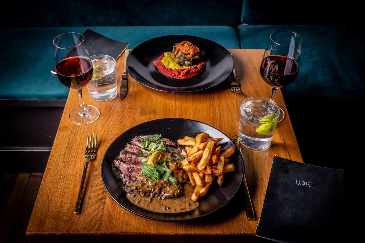 A wooden table set for two features plates of steak with fries and sauce, a dish with meat on red puree, two glasses of red wine, two glasses of water with lime, and black menus labeled "LORE.