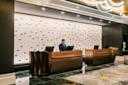 A person stands behind a modern hotel reception desk with two computer monitors. The lobby features elegant lighting, a patterned wall, and a decorative carpet.