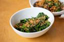 A white bowl filled with sautéed greens topped with crispy fried garlic sits on a wooden table, with another dish partially visible in the background.