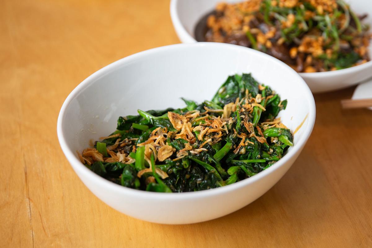 A white bowl filled with sautéed greens topped with crispy fried garlic sits on a wooden table, with another dish partially visible in the background.