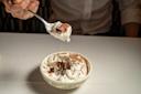 A person holds a spoonful of chocolate mousse topped with whipped cream above a bowl filled with more of the dessert, garnished with chocolate shavings, on a white tablecloth.
